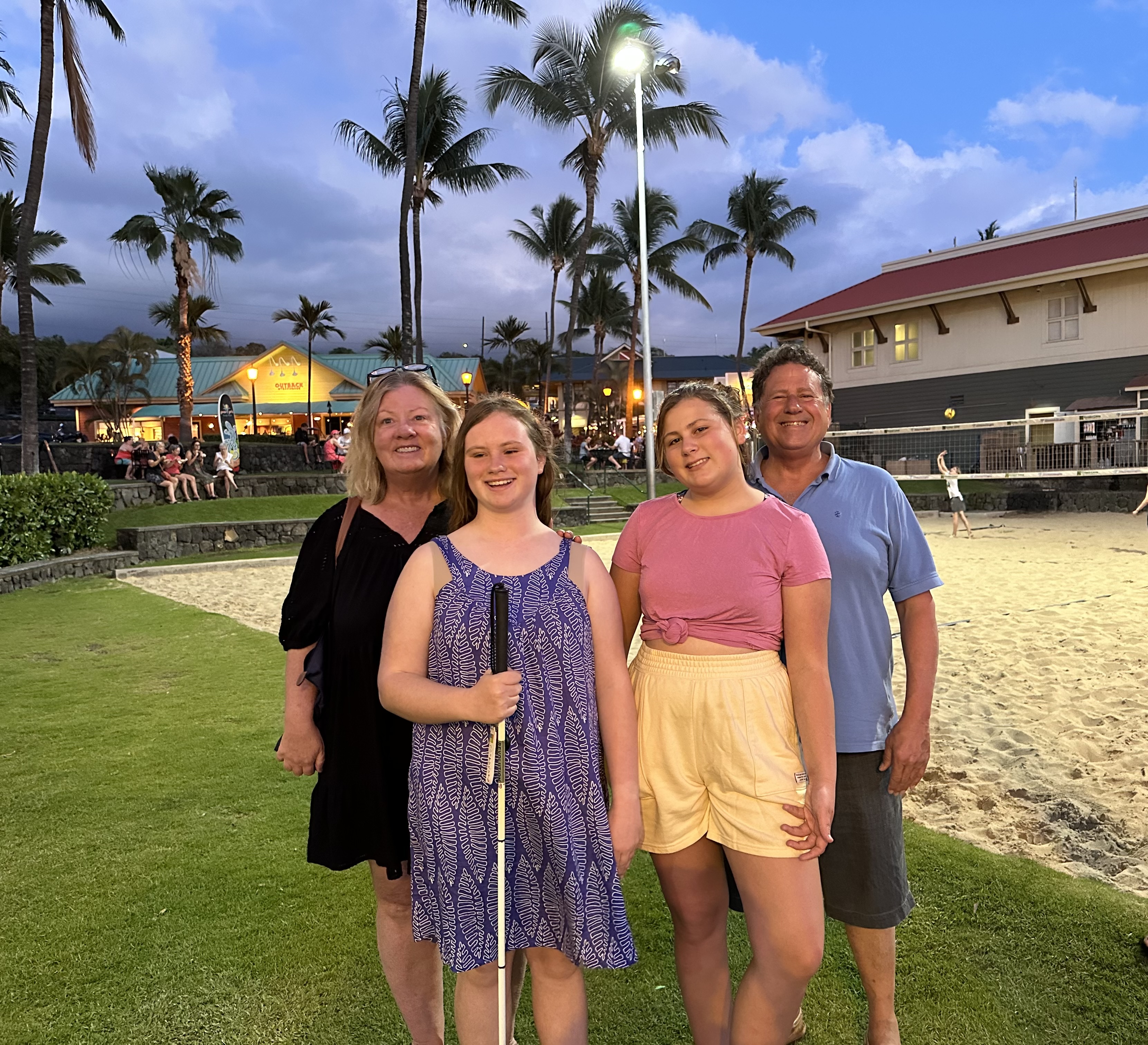 This picture shows the 4 members of the Dabbieri family standing on a grassy area near a beach in Hawaii. Julie Dabbieri is on the far left, wairing a black dress and has shoulder length blond hair. Grace Dabbieri is next to her, wairing a casual blue and white dress and holding a white cane for the blind. She has shoulder length brownish blond hair. The younger girl next to Grace, named Hope is wairing a pink top and yellow shorts. She also has shoulder length blond hair. The man on the right, Jon, is wairing a light blue shirt, and grey shorts. He has short brown hair. Behind them, there are tall palm trees, a sandy beech, and buildings with people arround. The sky above is a pretty blue and pink, indocating its evening time.